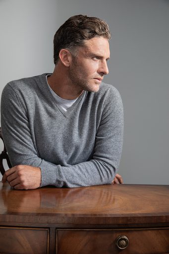 Johnstons of Elgin's Granite V Neck Cashmere Jumper on model wearing white t-shirt underneath leaning on wooden table
